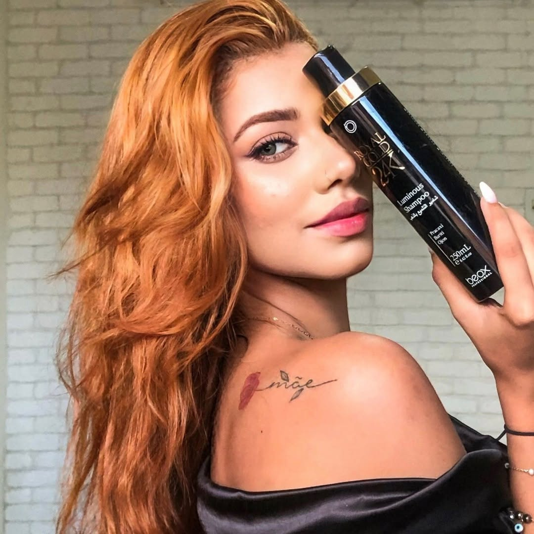 Woman with long red hair holding a bottle of Ouidad hair product against a brick wall.